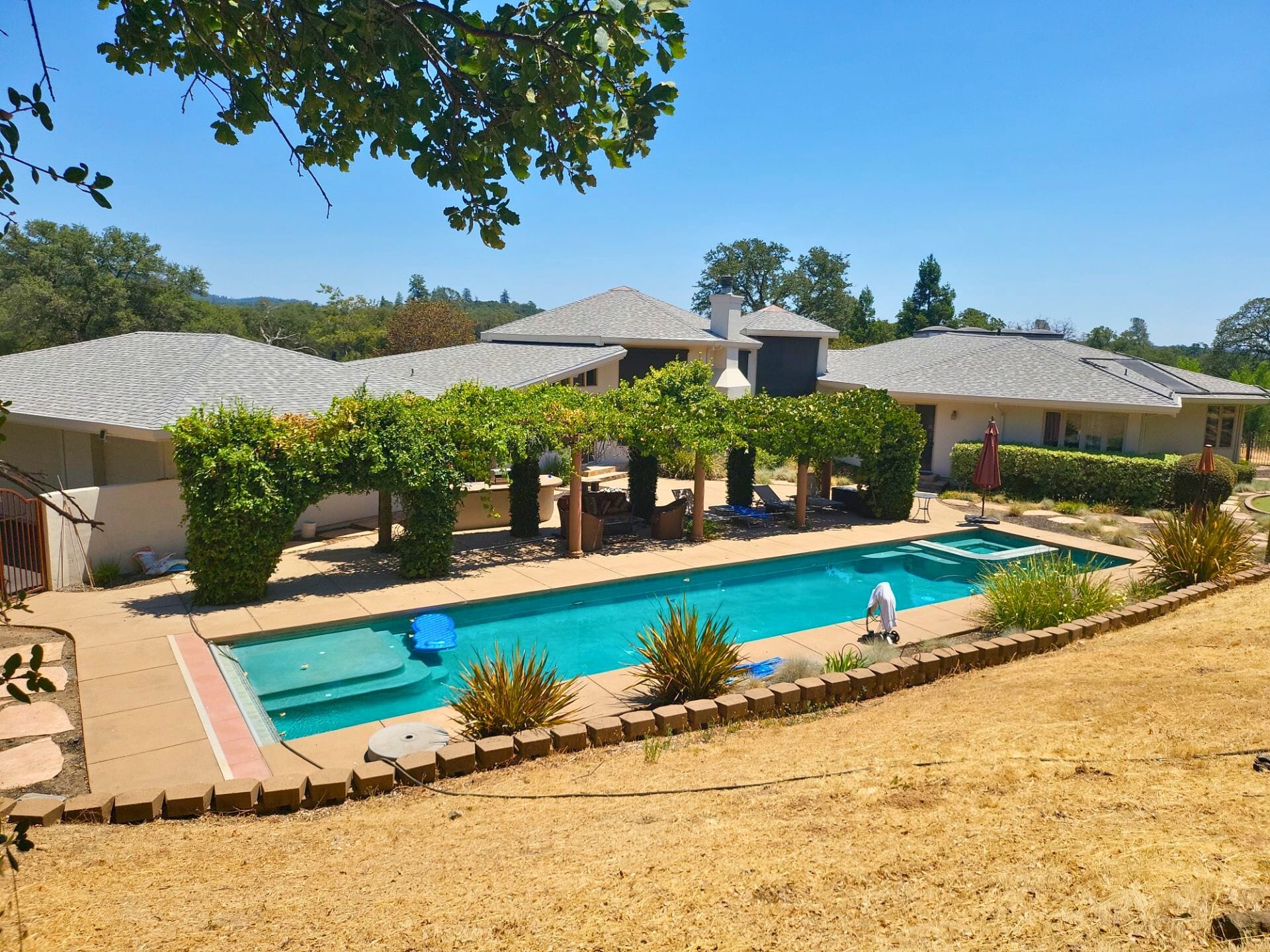 Before: angle 2. A backyard poolside view featuring a vine-covered pergola with wooden columns, outdoor furniture, and a built-in barbecue area, set against a white stucco house with a clear blue sky.