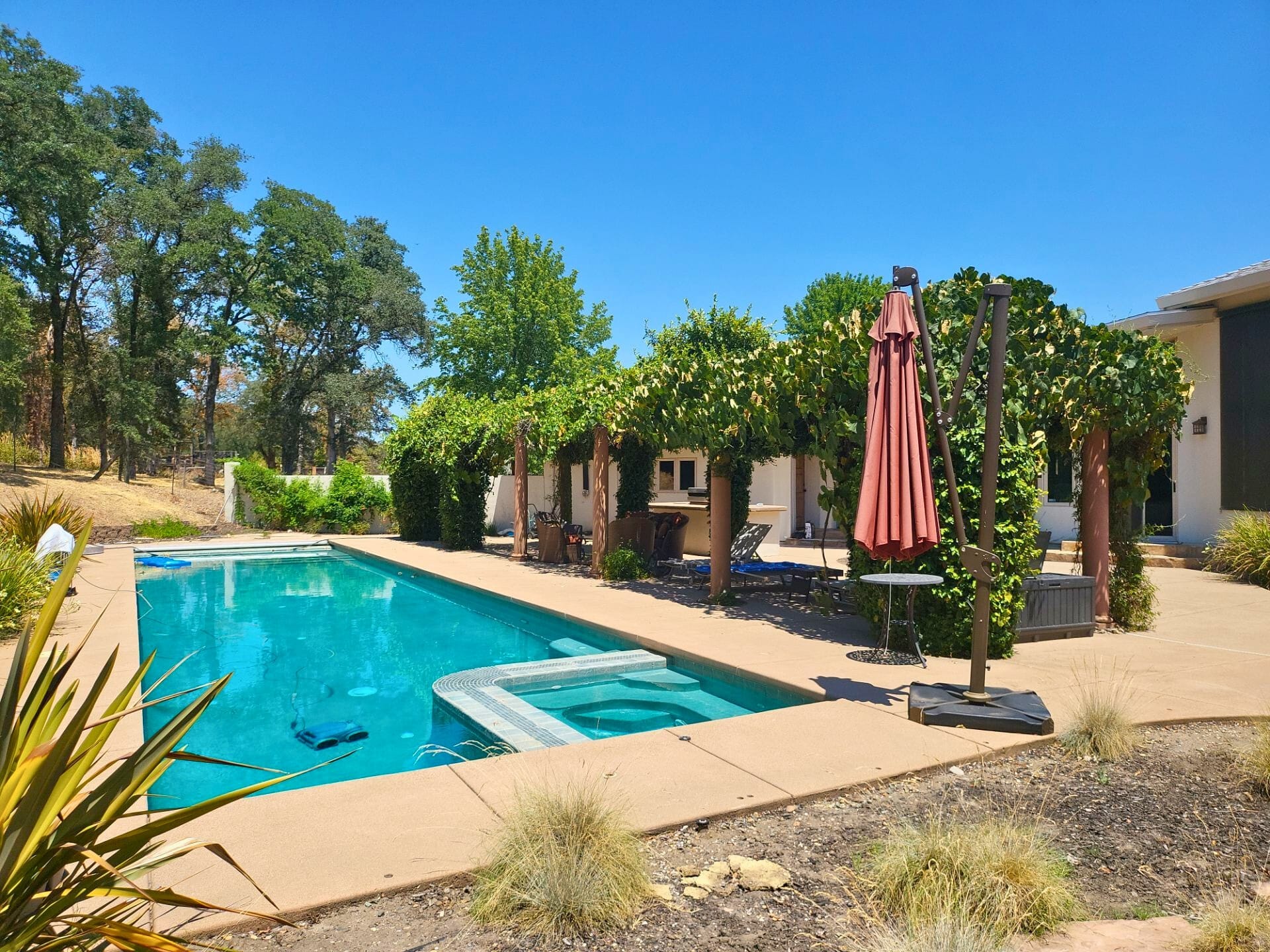 Before: angle 3. View featuring a vine-covered pergola with wooden columns, outdoor furniture, and a built-in barbecue area, set against a white stucco house with a clear blue sky.