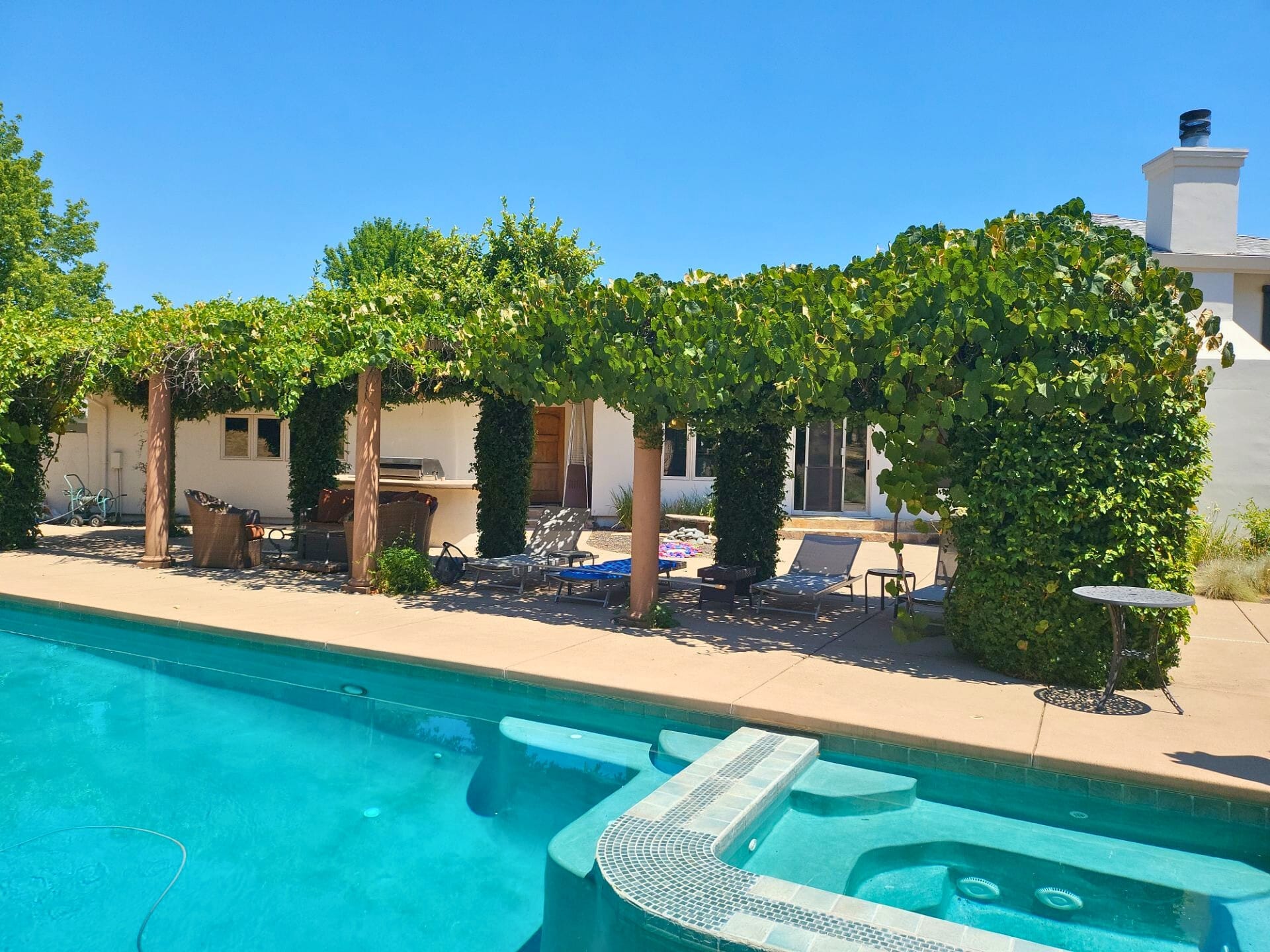 Before: angle 4. View featuring a vine-covered pergola with wooden columns, outdoor furniture, and a built-in barbecue area, set against a white stucco house with a clear blue sky.